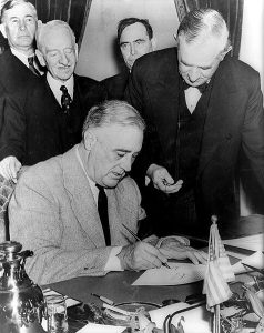 United States President Franklin D. Roosevelt signing the declaration of war against Germany, marking US entry into World War II in Europe. Senator Tom Connally stands by holding a watch to fix the exact time of the declaration. Date: 11 December 1941